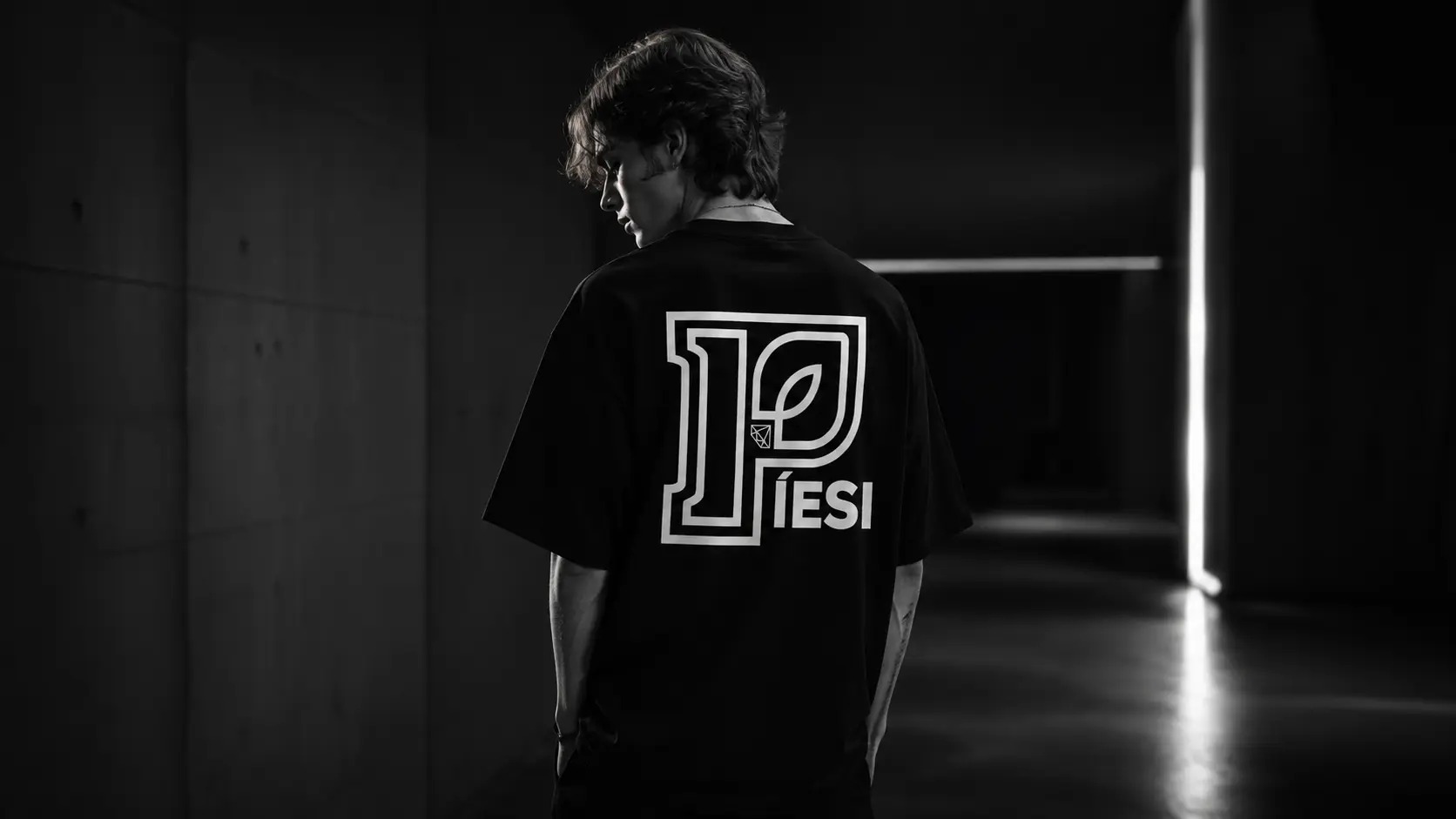 Back-view male model wearing the PÍESI Big P tee in a dark studio corridor.