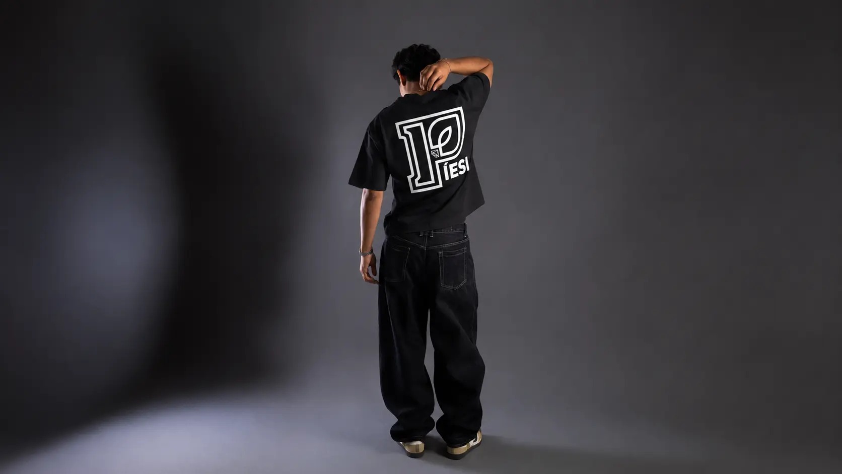 Back-view male model wearing the PÍESI Big P tee in a dark studio — editorial monochrome frame.