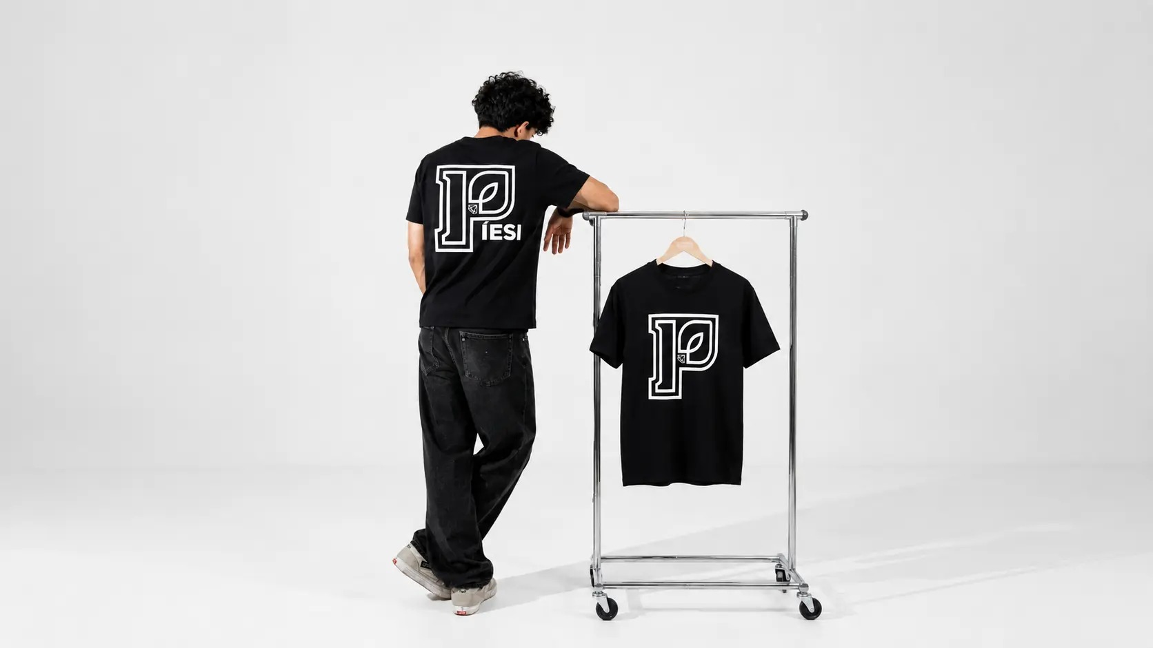 Back-view model with the PÍESI Big P tee next to a clothing rack on a white studio backdrop.